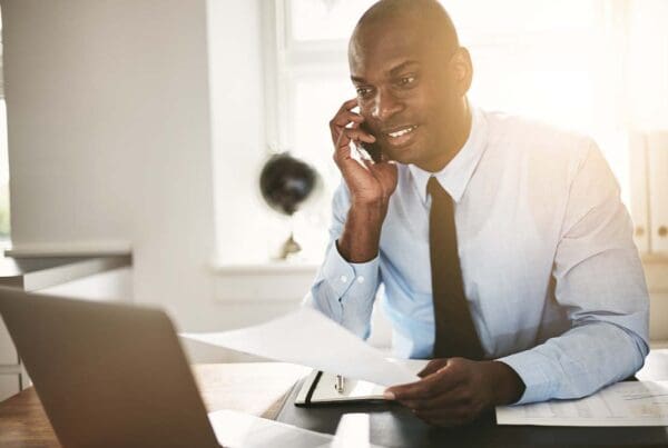 Surety Bond - Smiling Businessman Examining Documentation over the Phone and Working from His Home Office and Using a Laptop